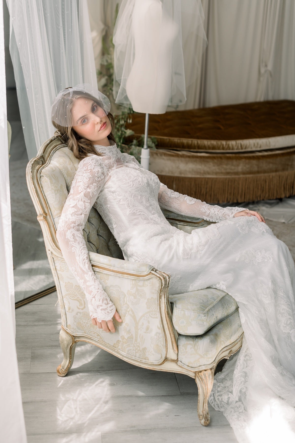 Woman in a white lace dress sitting on an ornate chair in a softly lit room.