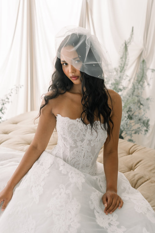 Woman in a white lace wedding dress with a veil sitting on a couch.
