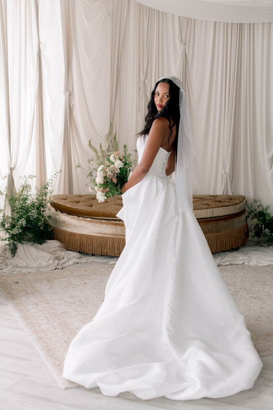 Woman in a white wedding dress standing in a decorated room with floral arrangements and a large woven basket.