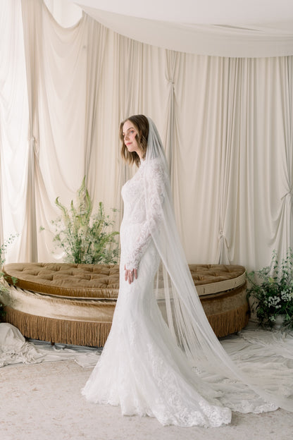 Woman in a white wedding dress with a long veil standing in a decorated room with curtains and plants.