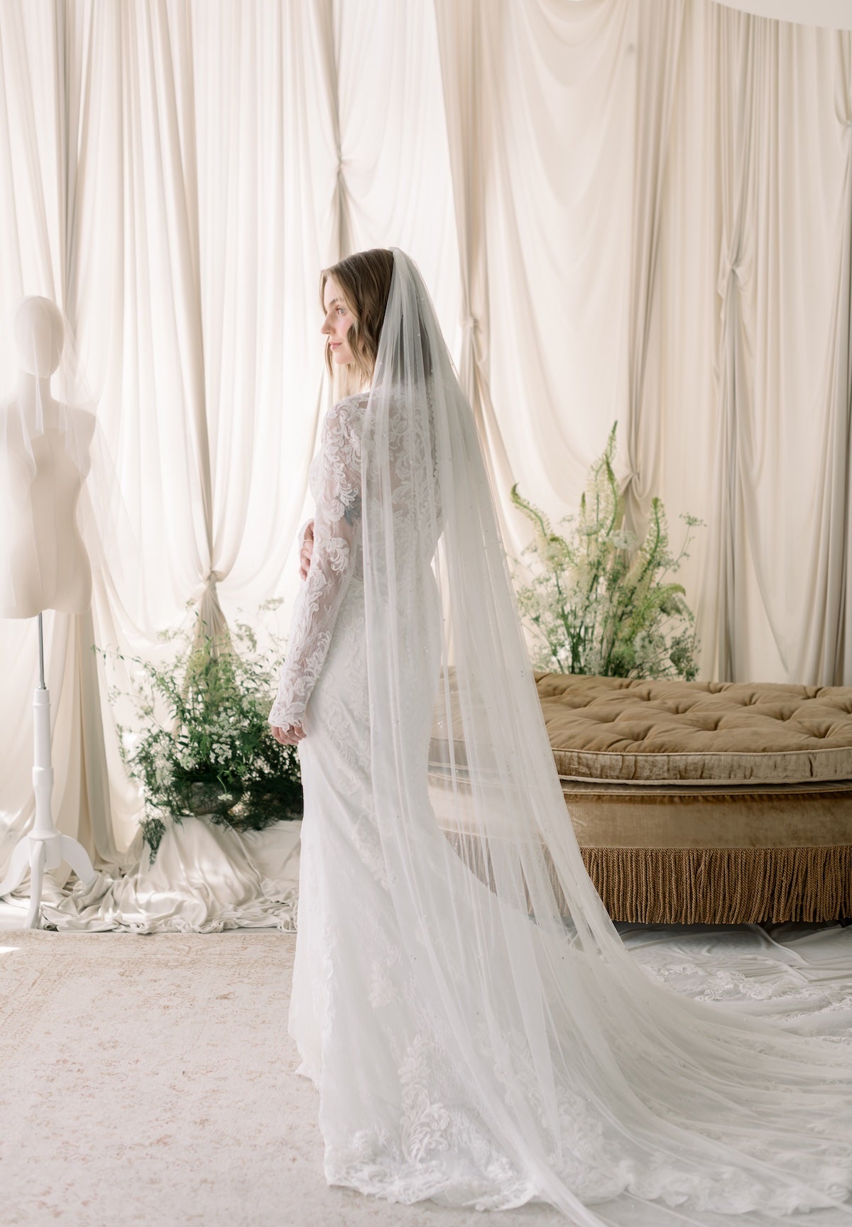 Woman in a wedding dress with a long veil standing in a room with white curtains and a beige couch.
