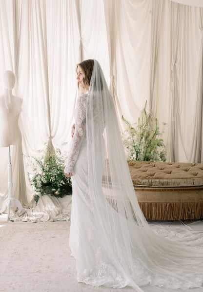 Woman in a wedding dress with a long veil standing in a room with white curtains and a beige couch.