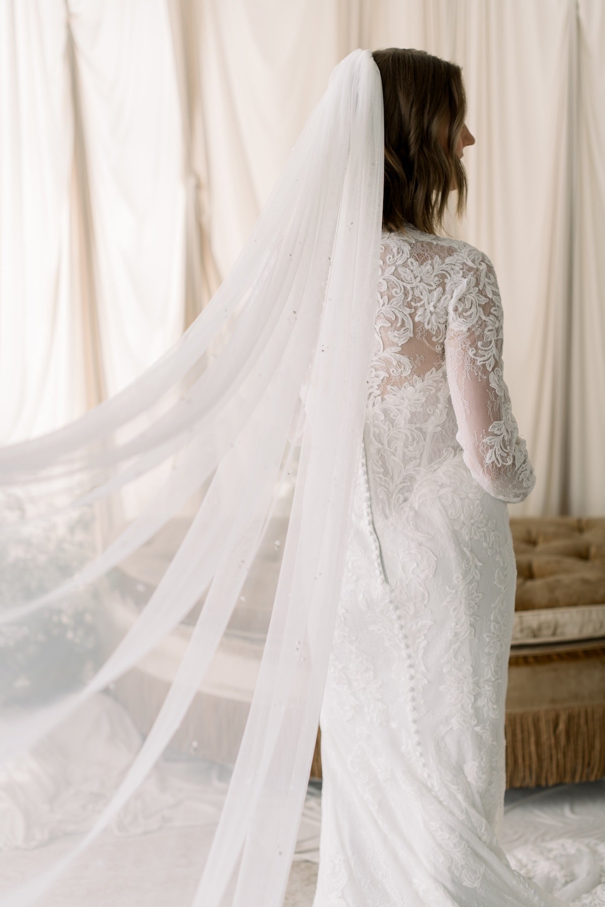 Woman wearing a lace wedding dress with a long veil in a softly lit room.