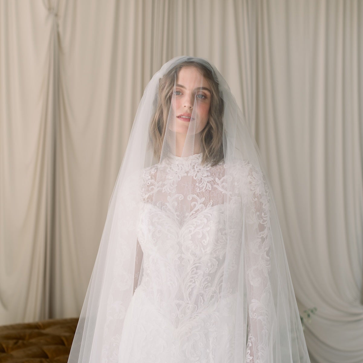 Woman wearing a white lace wedding dress with a matching veil against a neutral background