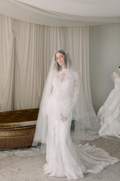 Woman wearing a white lace wedding dress with a long train in a room with curtains.