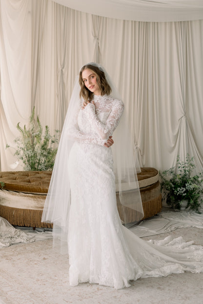 Woman in a white wedding dress standing in a softly lit room with draped curtains and floral decorations.