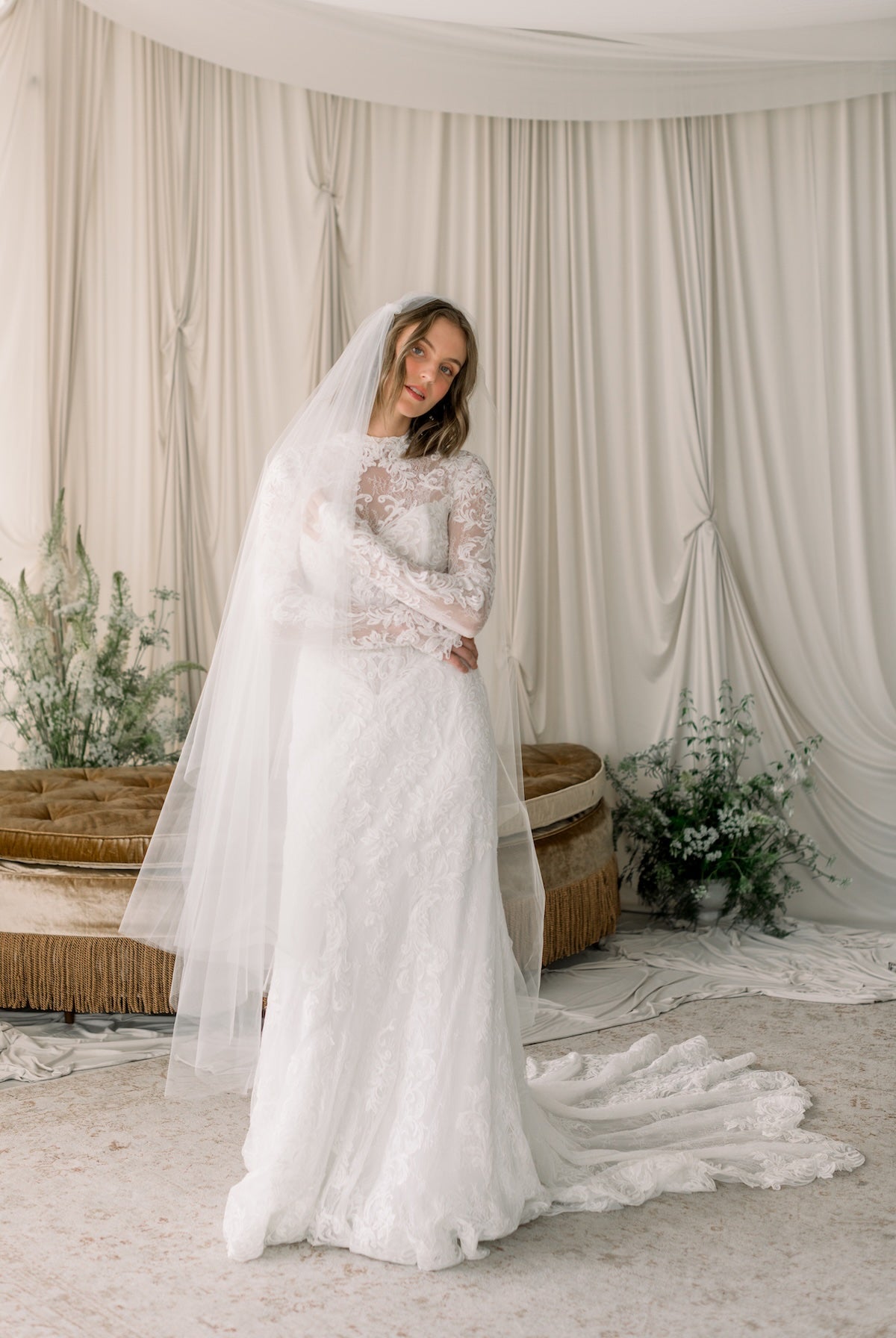 Woman in a white wedding dress with a long veil standing in a room with white curtains and decorative plants.