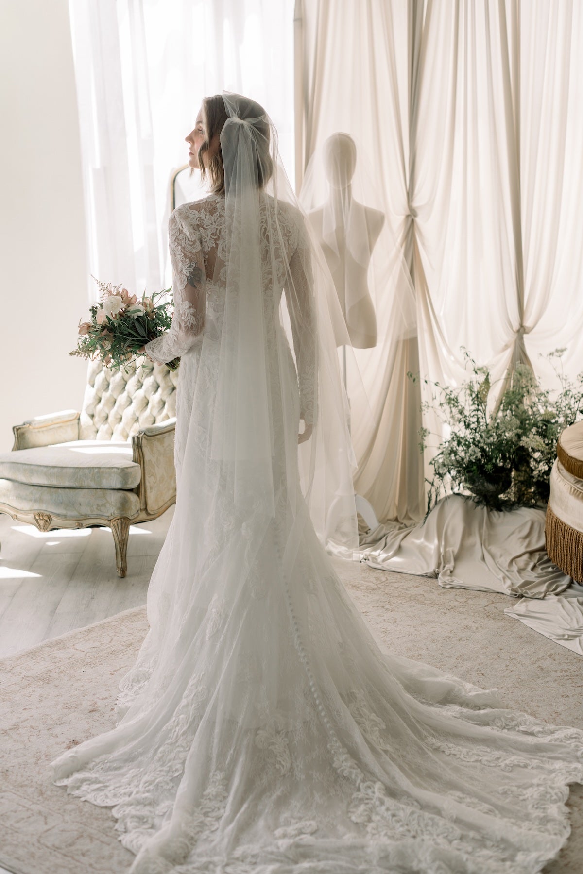 Bride in a white lace wedding dress standing in a elegant room with curtains and flowers.