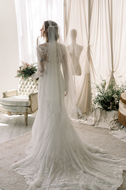 Bride in a white lace wedding dress standing in a elegant room with curtains and flowers.