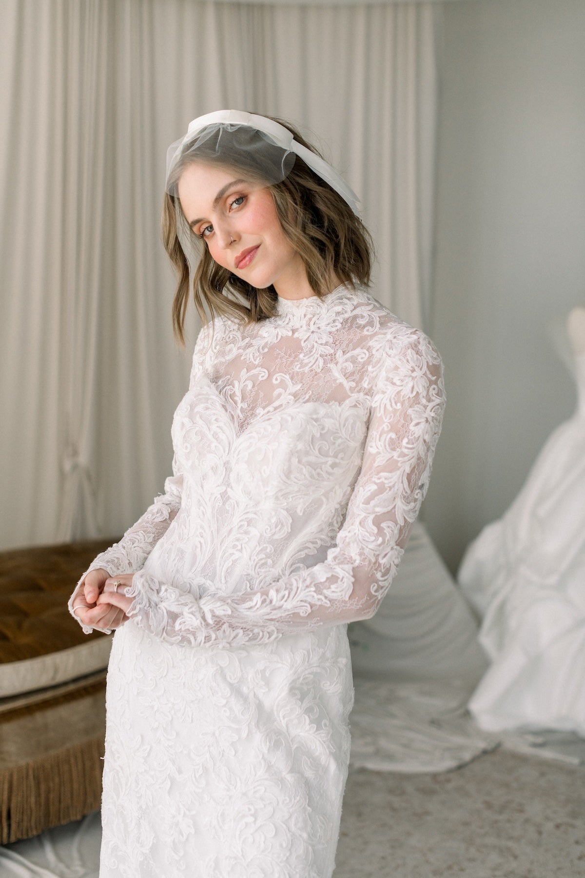 Woman wearing a lace wedding dress with a veil in a room with white curtains.