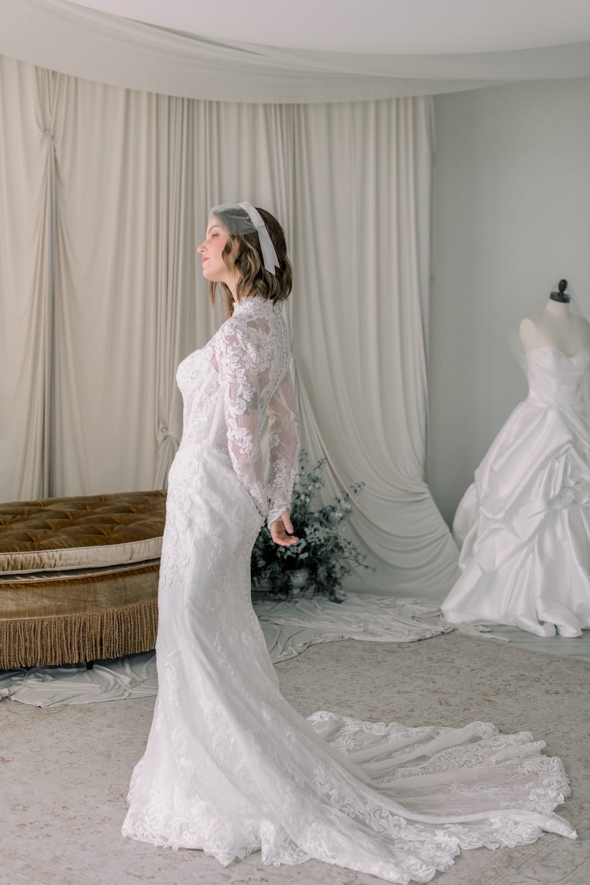 Woman in a white lace wedding dress standing in a room with draped curtains and another wedding dress.