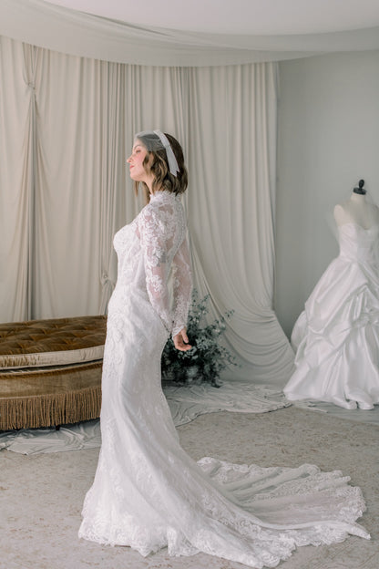 Woman in a white lace wedding dress standing in a room with draped curtains and another wedding dress.