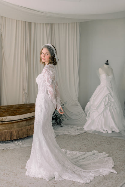 Woman in a white lace wedding dress standing in a room with draped curtains and another wedding dress on a mannequin.