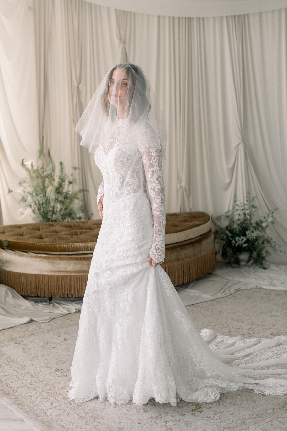 Bride in a white lace wedding dress with long sleeves and a veil, standing in a decorated room.