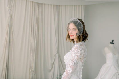 Woman wearing a lace wedding dress with a veil in a room with white curtains.