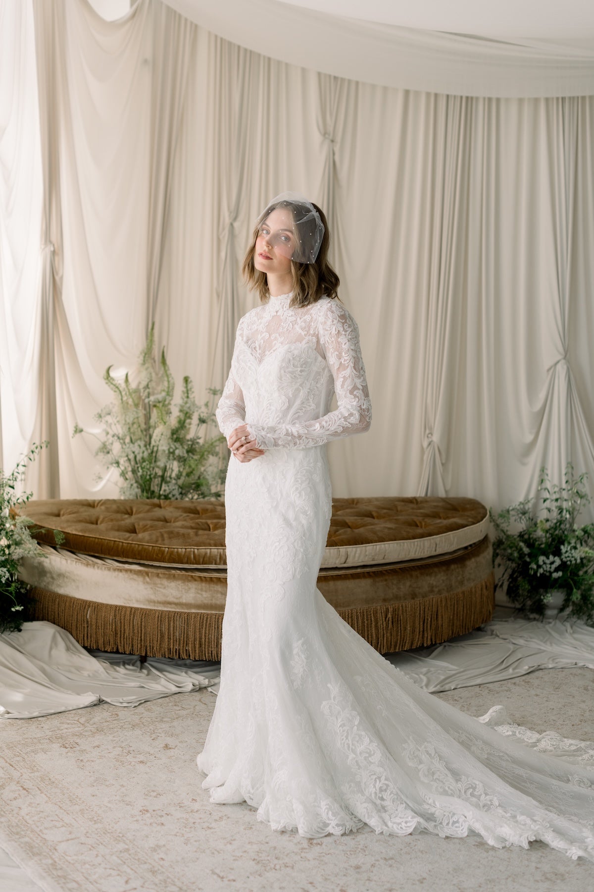 Woman in a white lace wedding dress standing in a decorated room with curtains and plants.