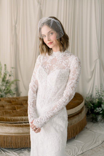 Woman wearing a white lace dress with a sheer veil in a softly lit room.
