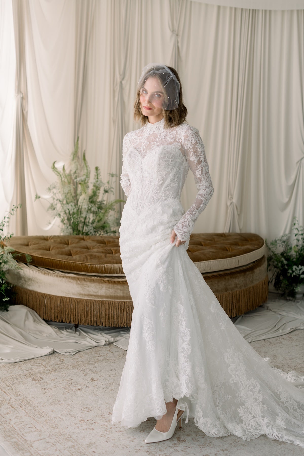 Woman in a lace wedding dress standing in a decorated room with curtains and a couch.