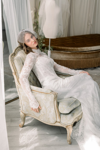 Woman in a white lace dress sitting on an ornate chair in a softly lit room.