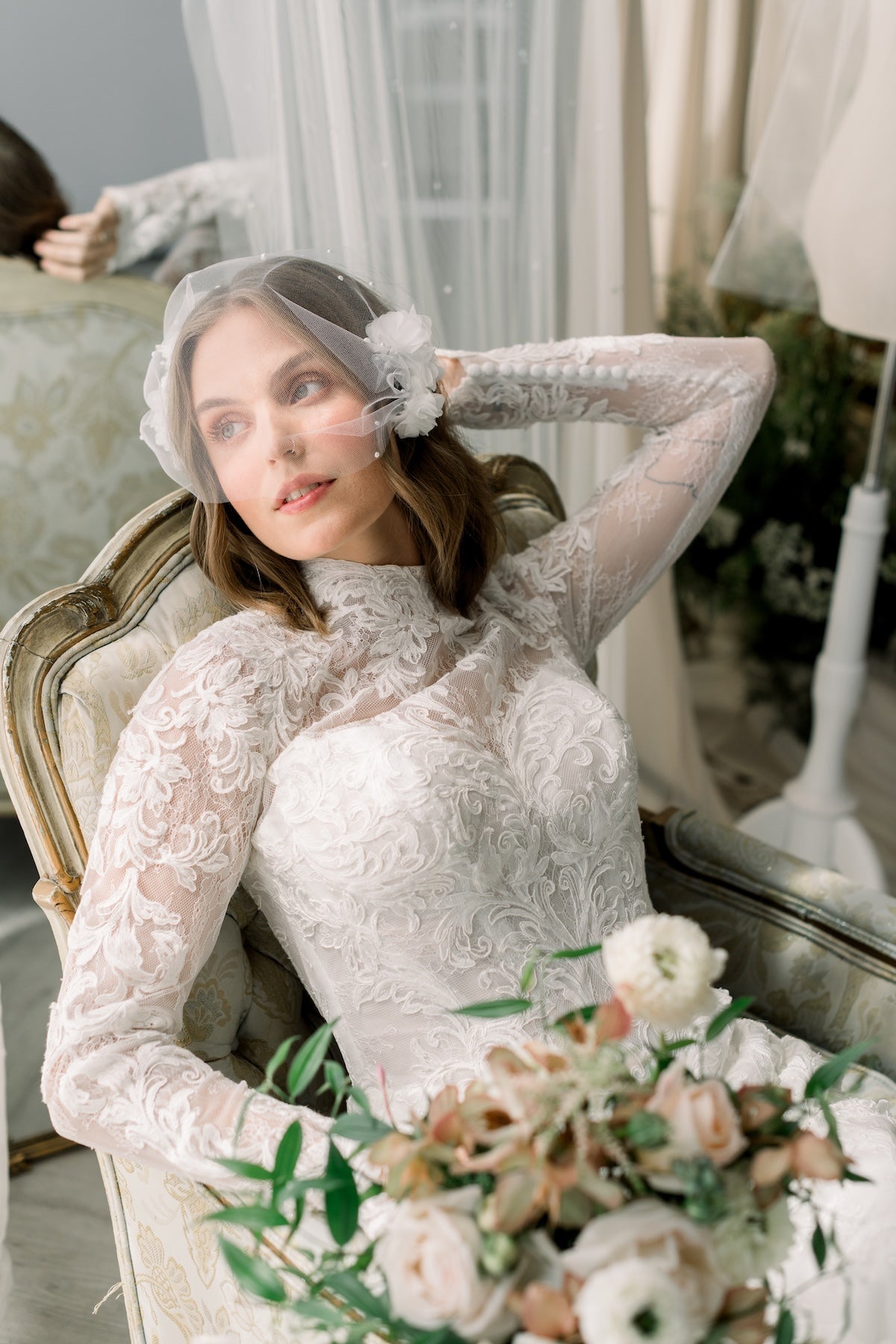 Woman in a lace wedding dress sitting on a chair with a bouquet of flowers, wearing a veil.