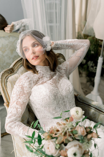 Woman in a lace wedding dress sitting on a chair with a bouquet of flowers, wearing a veil.