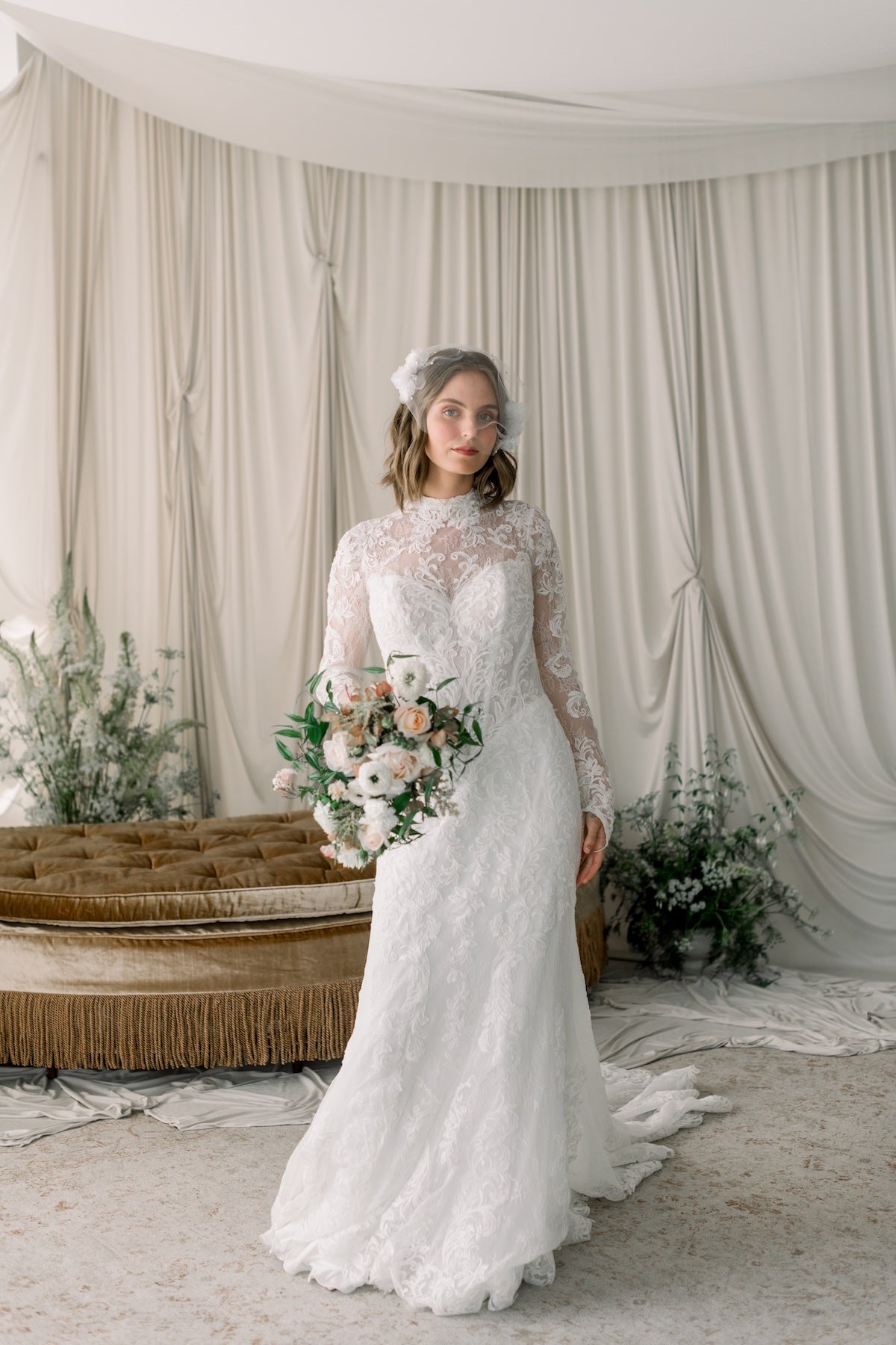 Woman in a white lace wedding dress holding a bouquet in a decorated room with draped curtains.