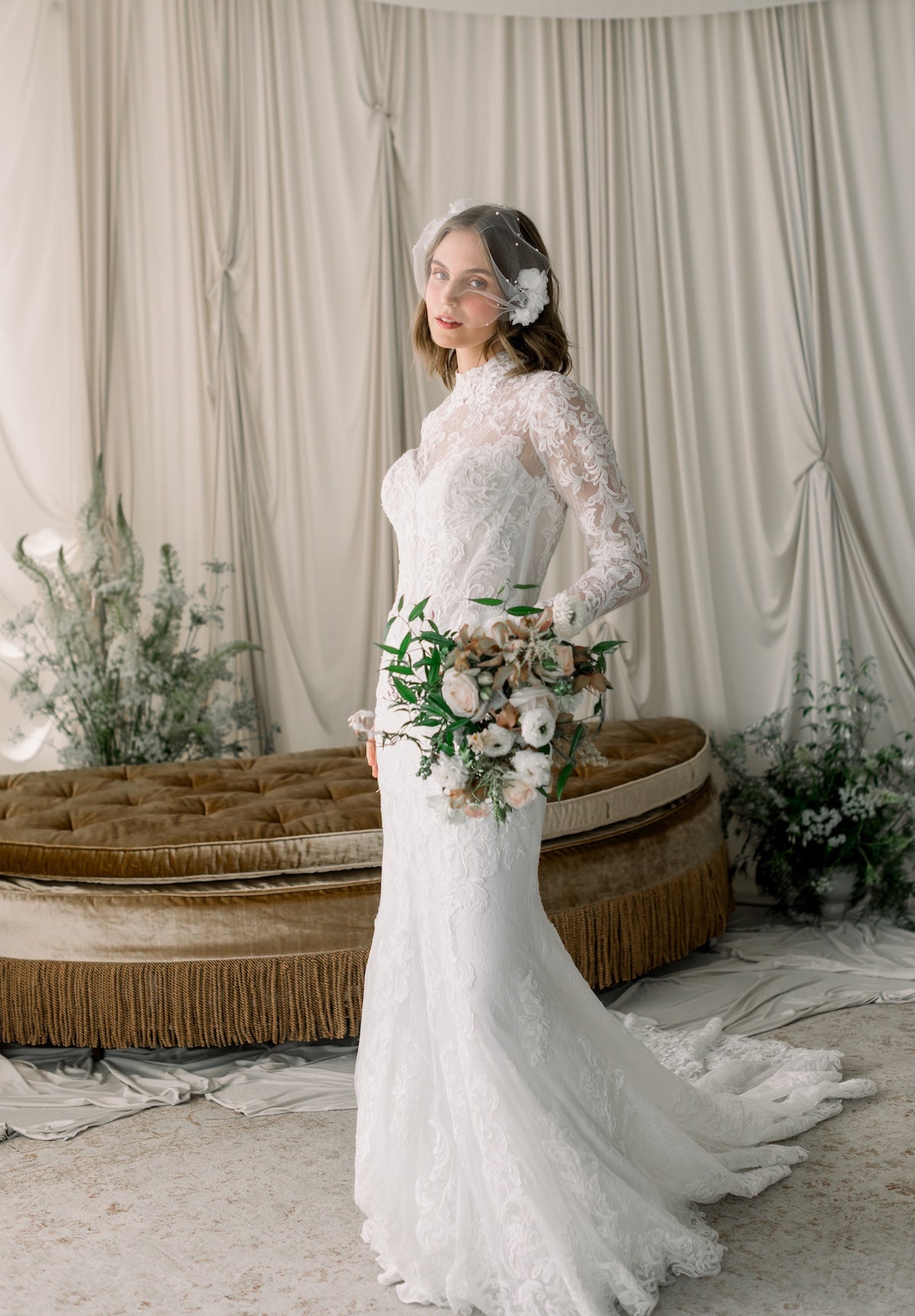 Woman in a white lace wedding dress holding a bouquet in a decorated room.
