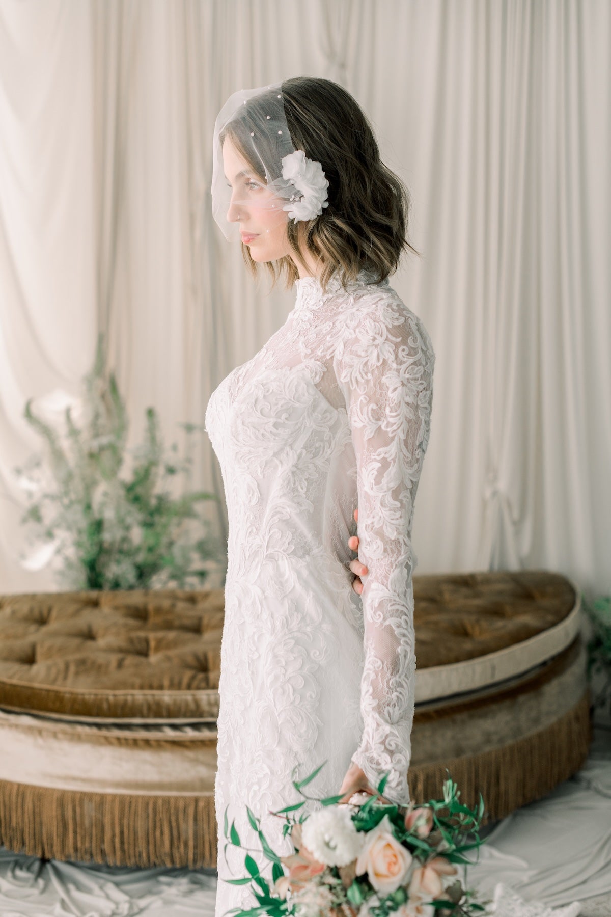 Woman in a white lace dress holding a bouquet, wearing a floral headband in a softly lit room.