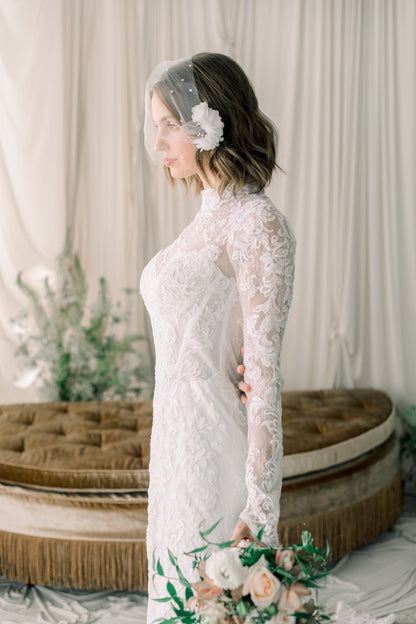 Woman in a white lace dress holding a bouquet, wearing a floral headband in a softly lit room.