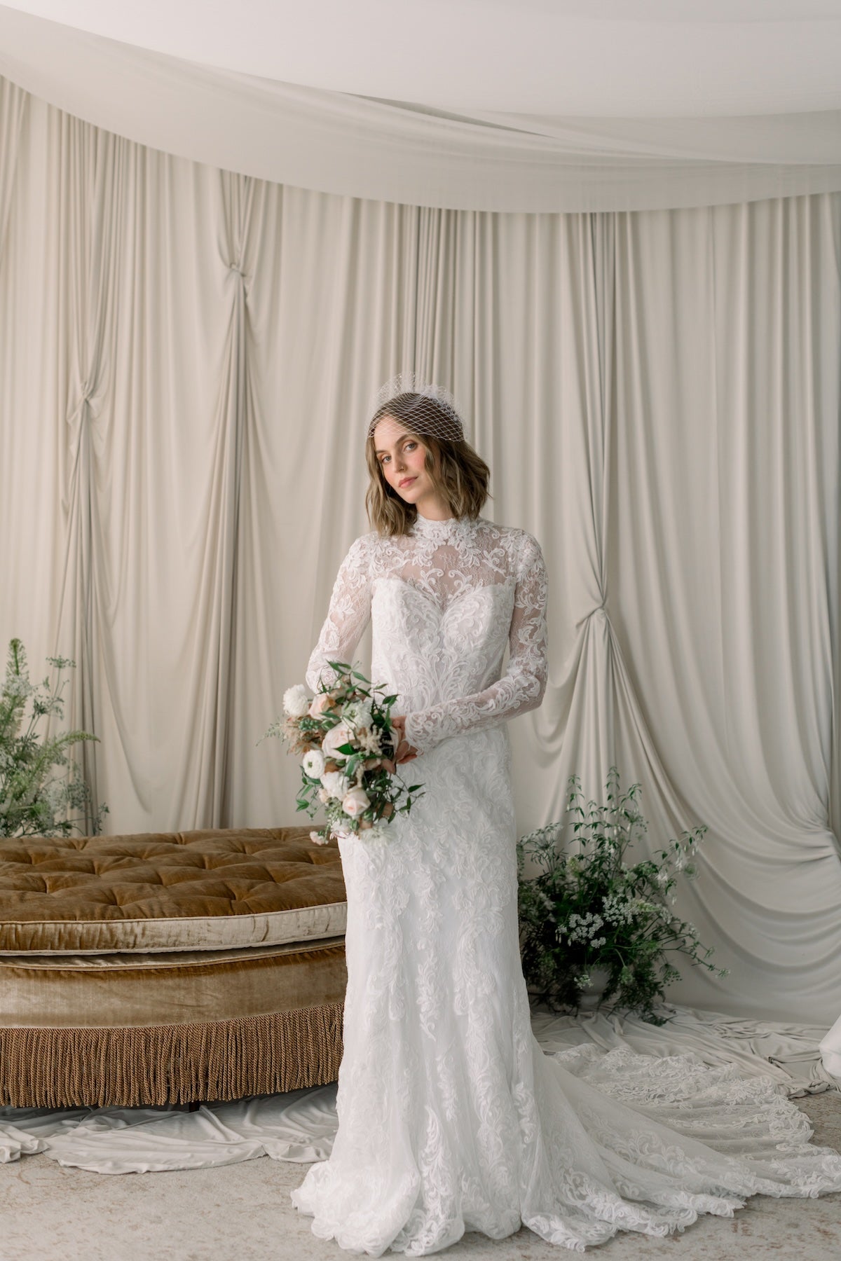 Woman in a lace wedding dress holding flowers in a decorated room with curtains and a couch.