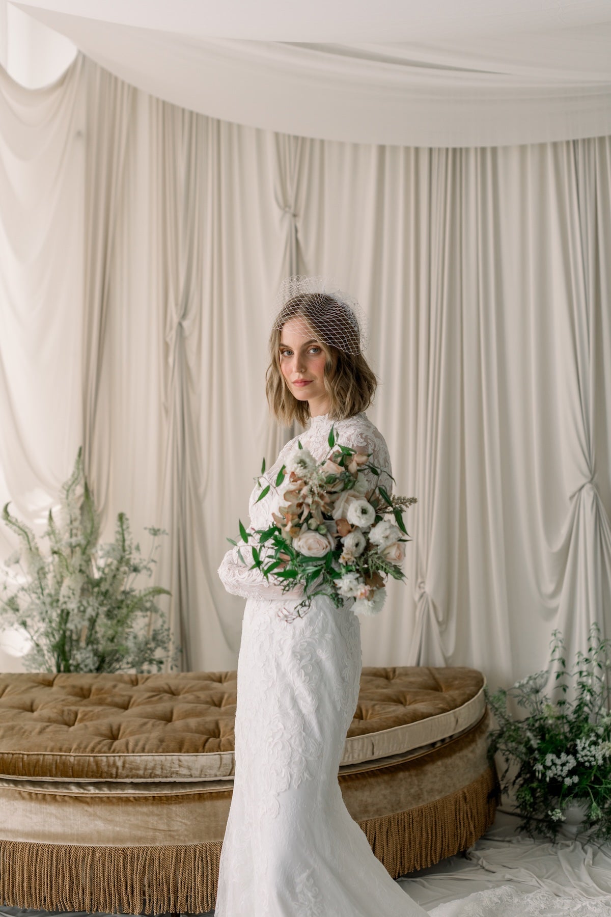 Woman in a white dress holding a bouquet in a decorated room with curtains and flowers.