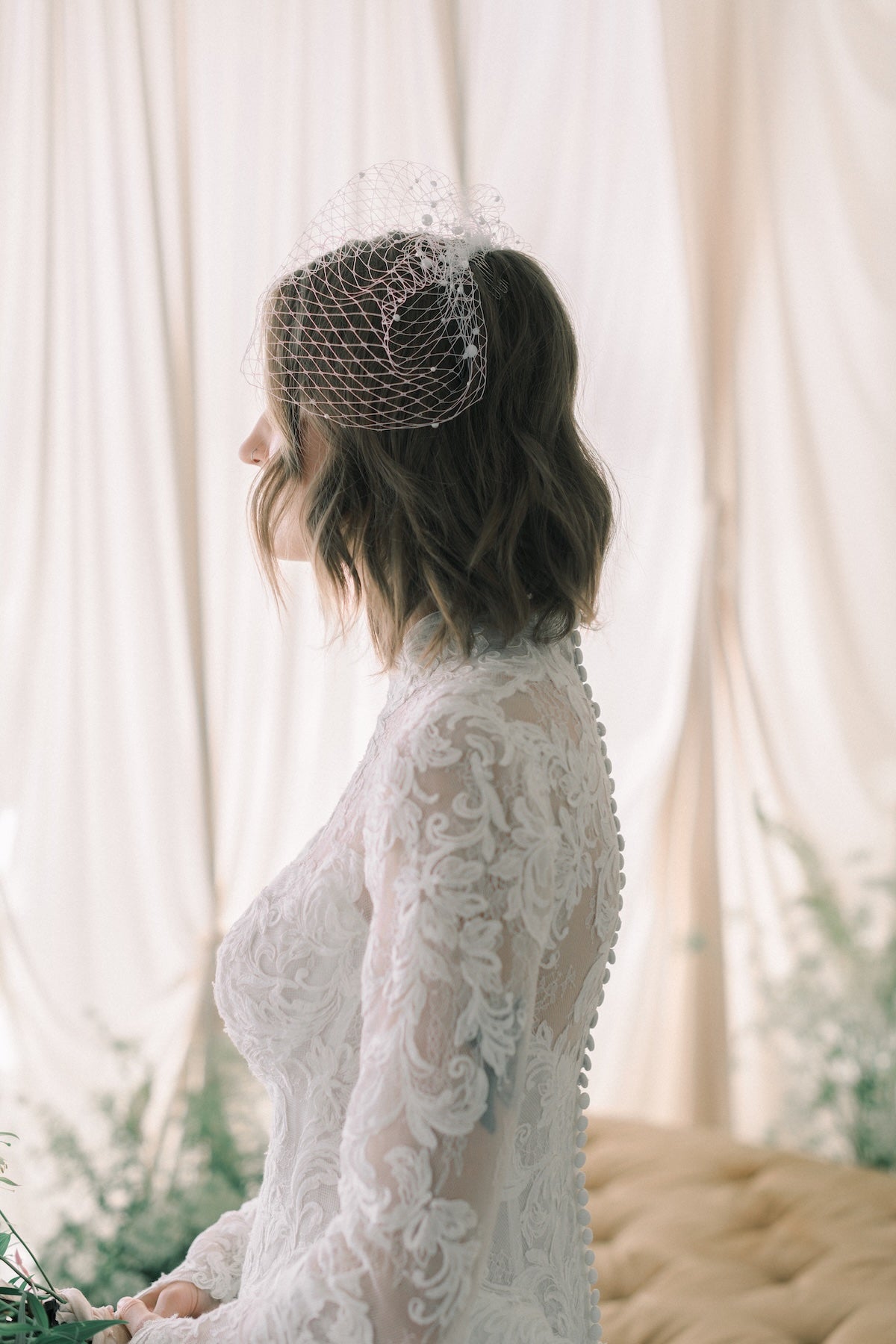 Woman in a white lace dress with a birdcage veil against a white curtain background