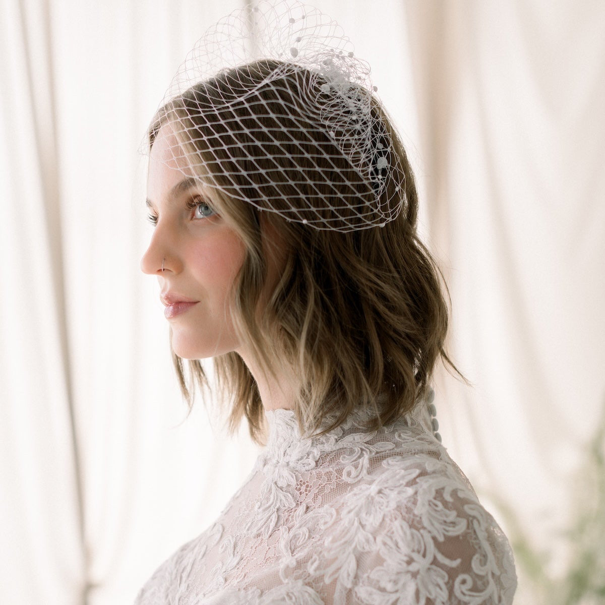 Woman wearing a lace wedding dress and birdcage veil against a soft, blurred background