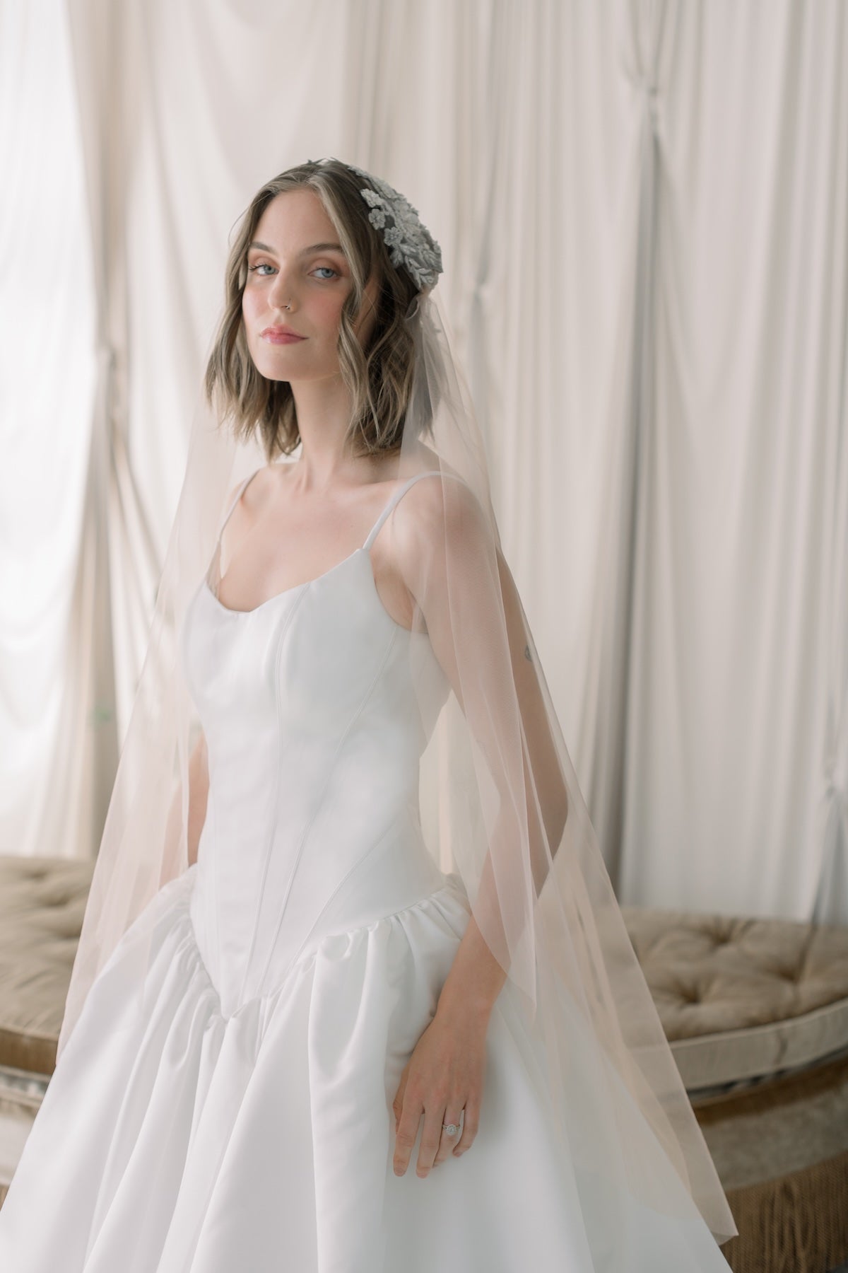 Woman in a white wedding dress with a sheer overlay and floral headpiece against a soft, neutral background.