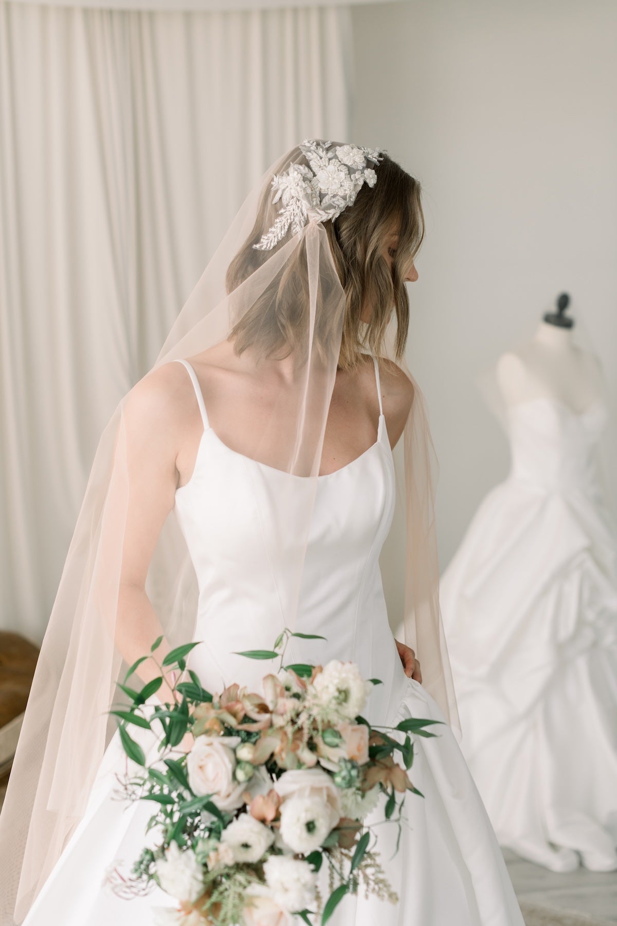 Bride in a white wedding dress with a floral headpiece and bouquet, standing in a room with a mannequin and curtains.