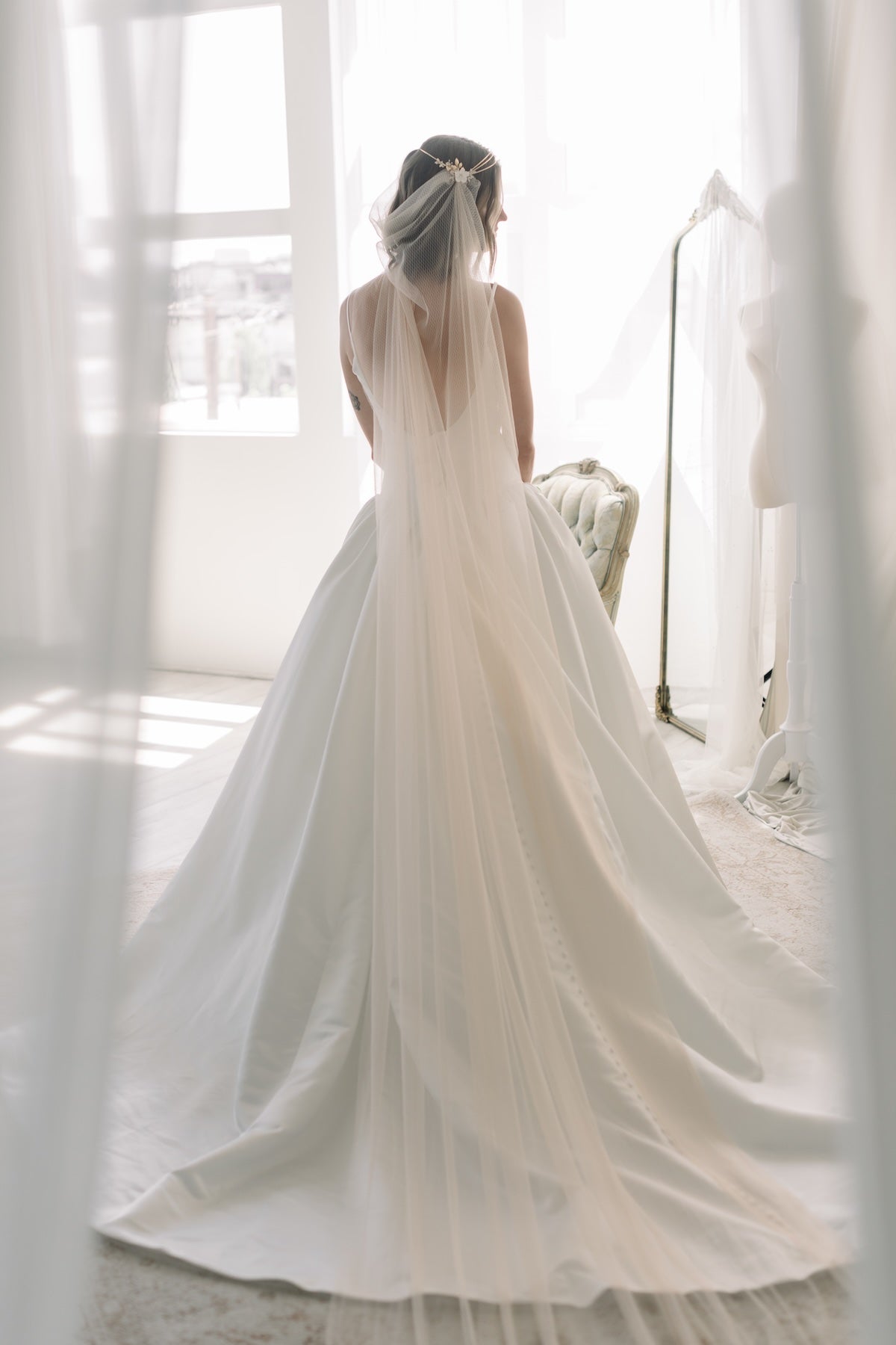Woman in a white wedding dress with a long train, standing in a softly lit room.