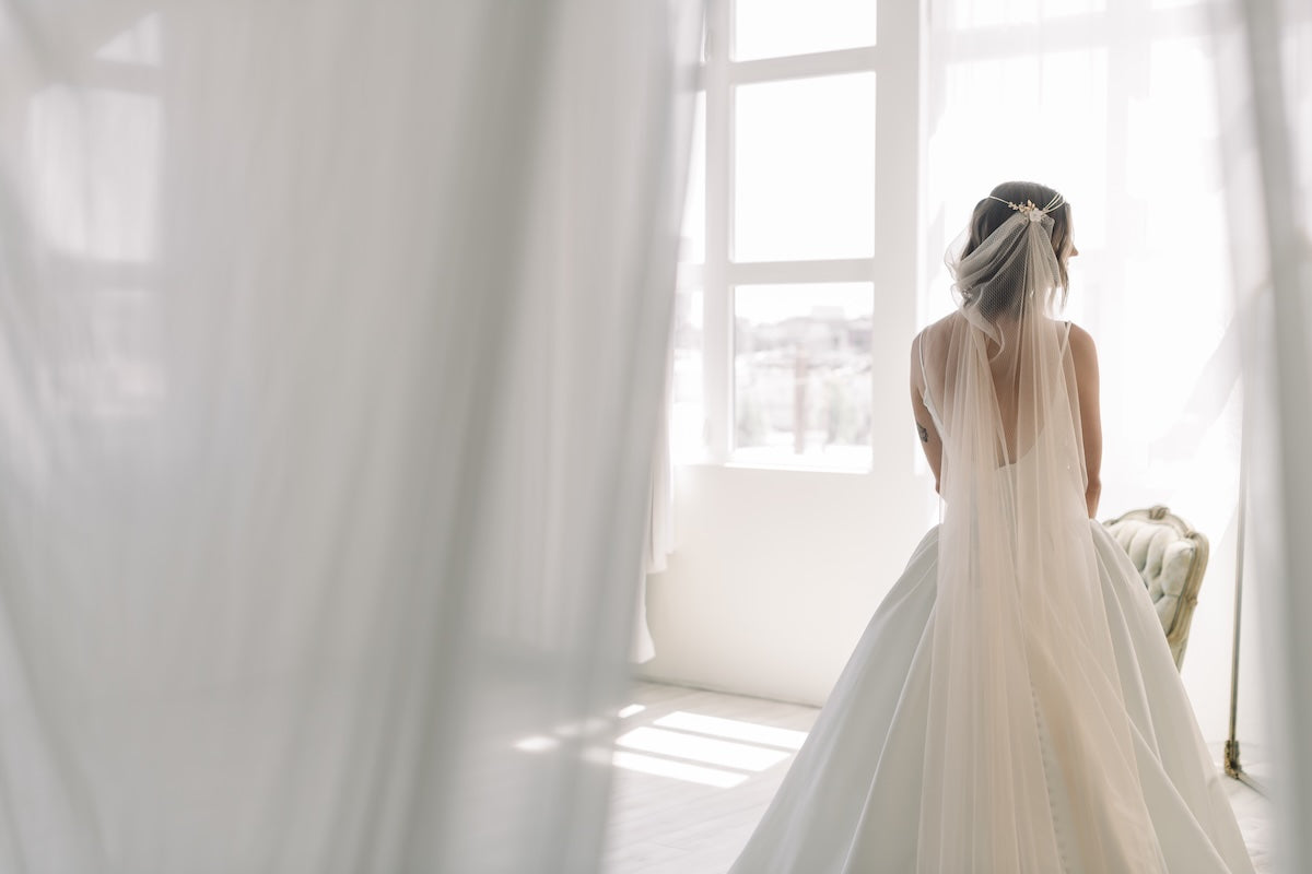 Woman in a wedding dress standing in a bright room with sheer curtains.