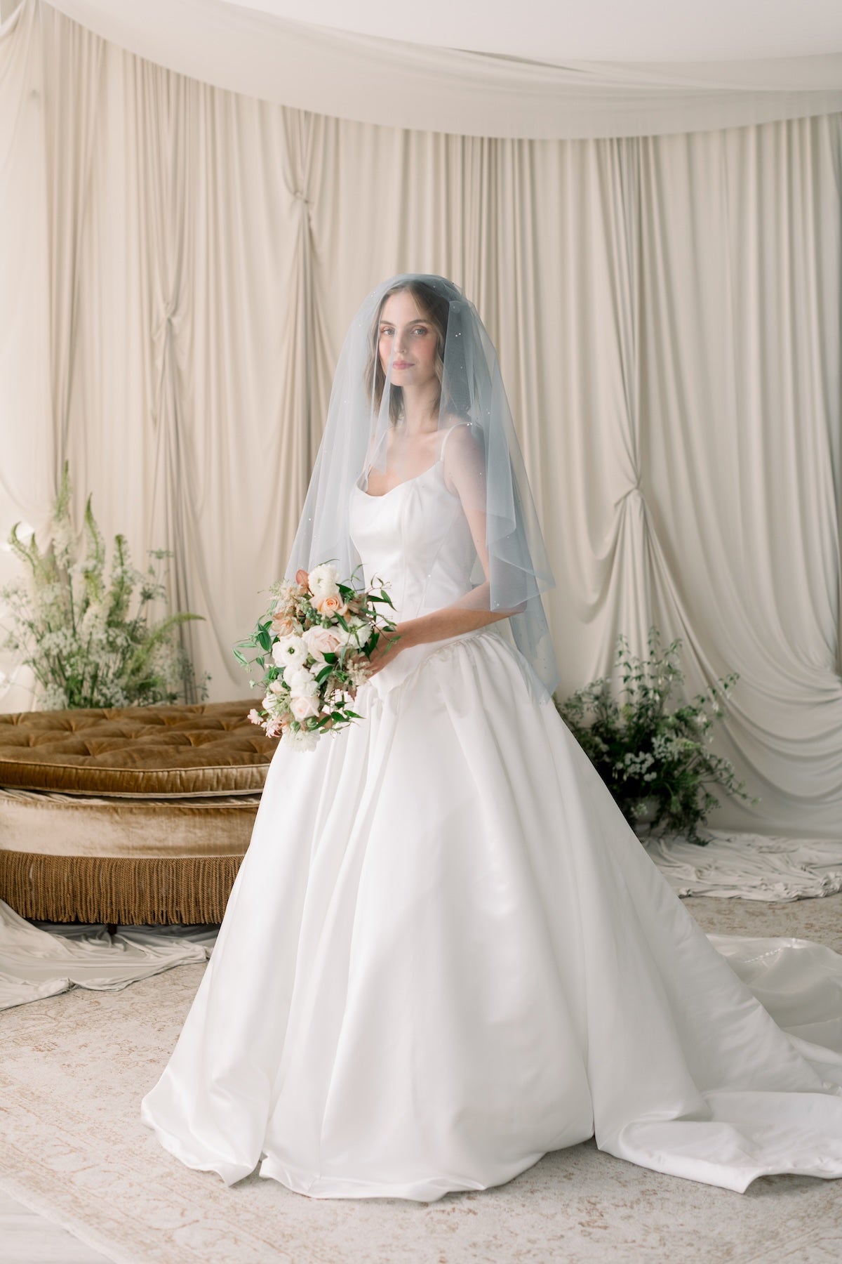Bride in a white wedding dress holding a bouquet in a decorated room with curtains and plants.