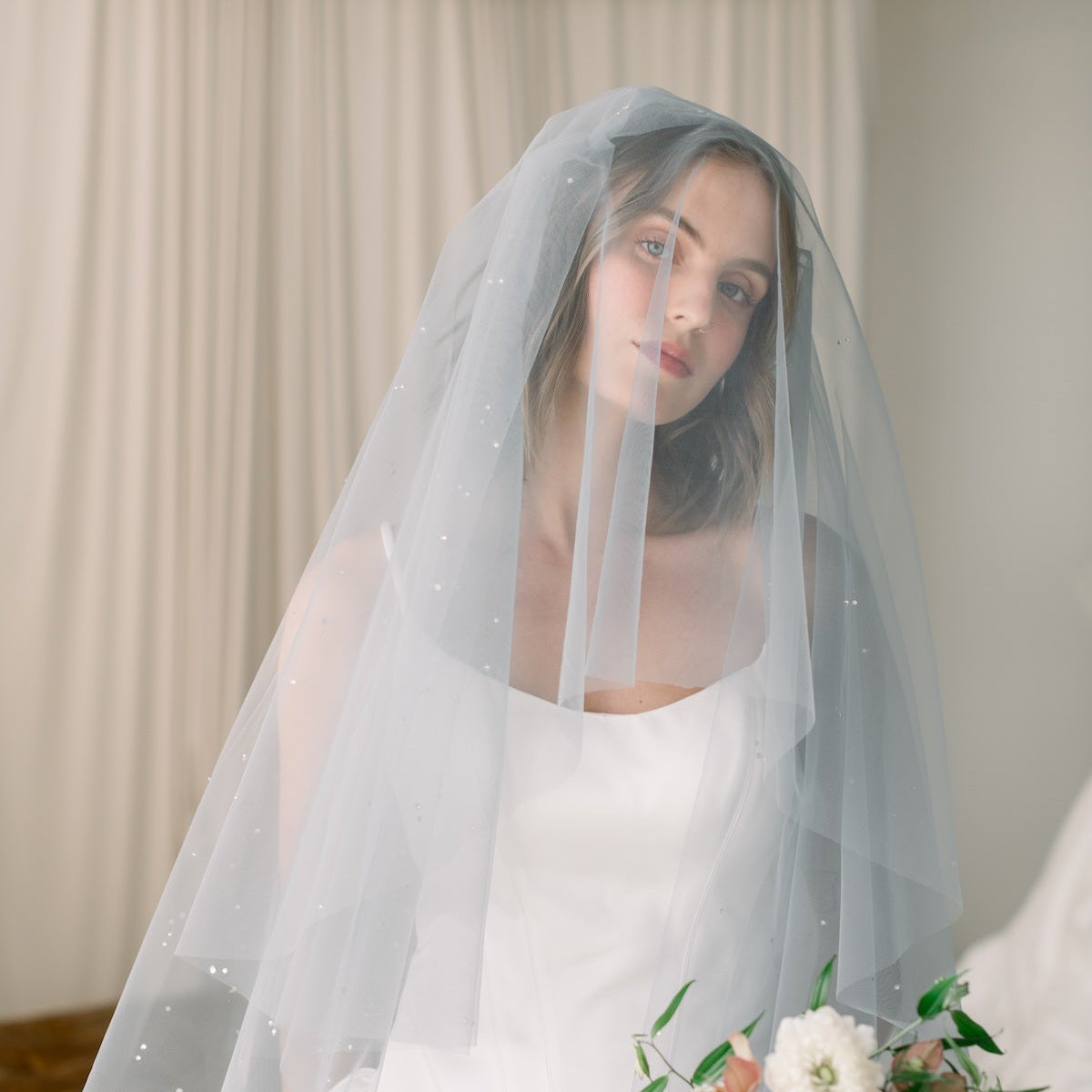 Bride in a white wedding dress with a veil holding a bouquet indoors.