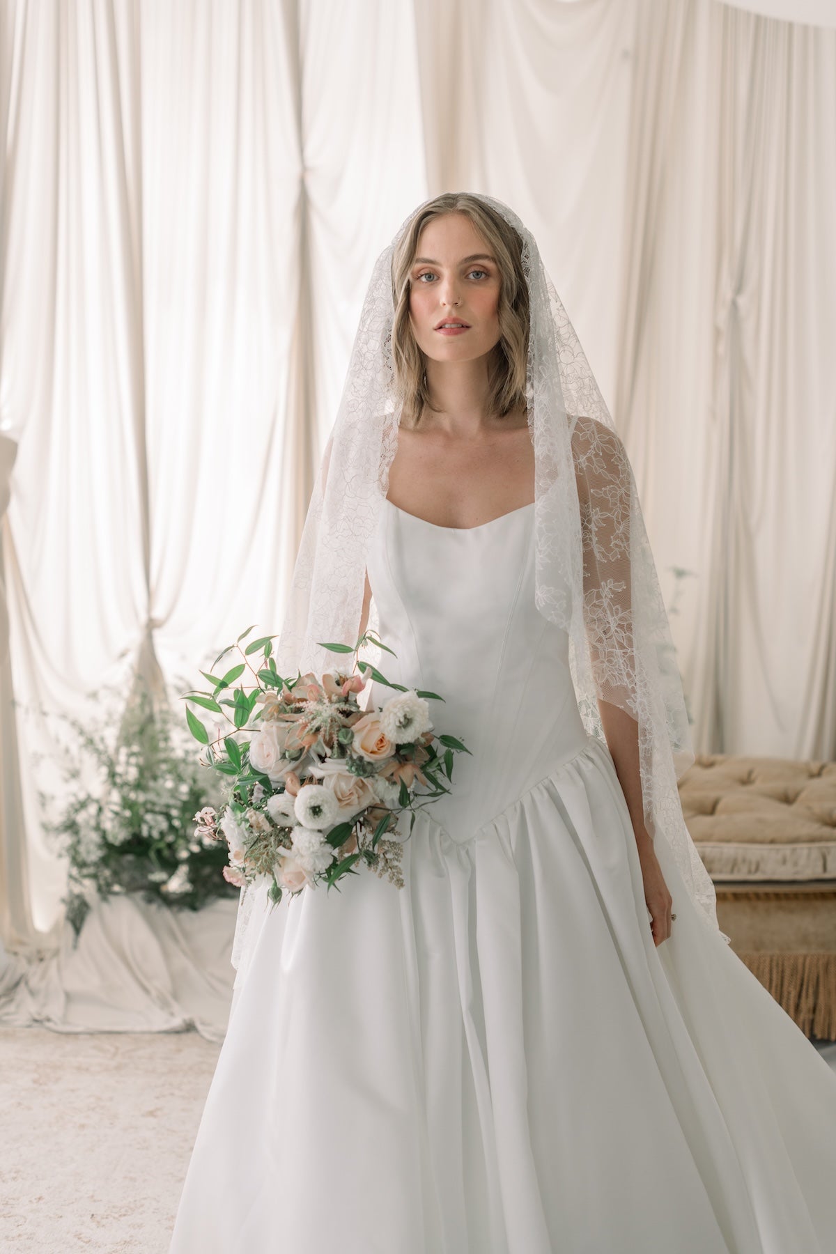 Bride in a white wedding dress with a veil holding flowers in a softly lit room.