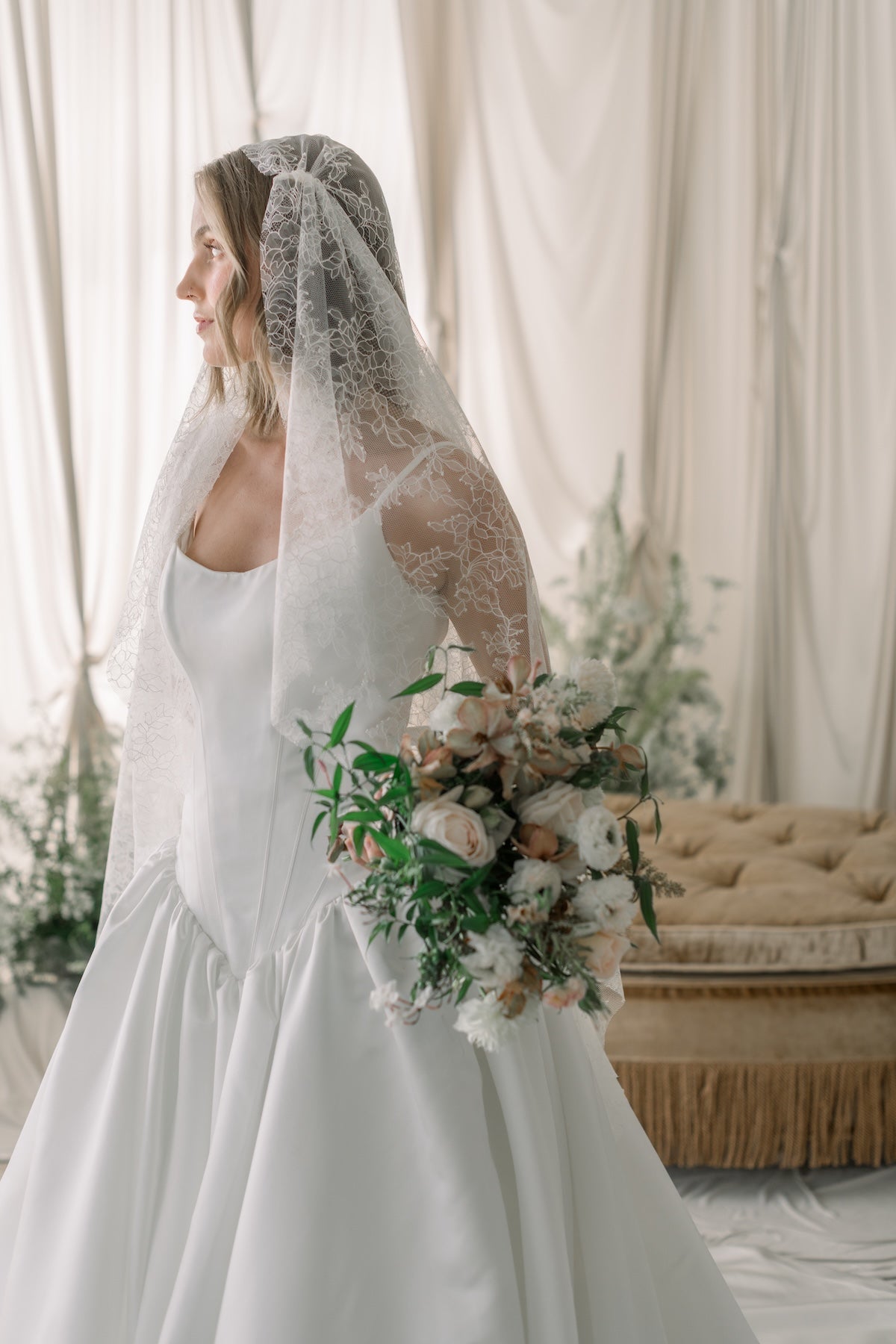 Bride in a white dress holding a bouquet with a lace veil, standing in a softly lit room.