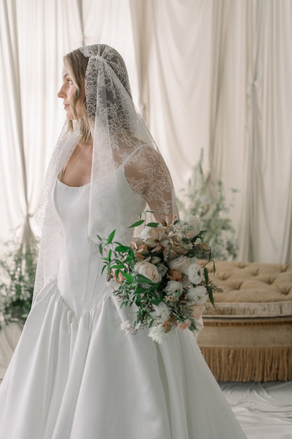 Bride in a white dress holding a bouquet with a lace veil, standing in a softly lit room.