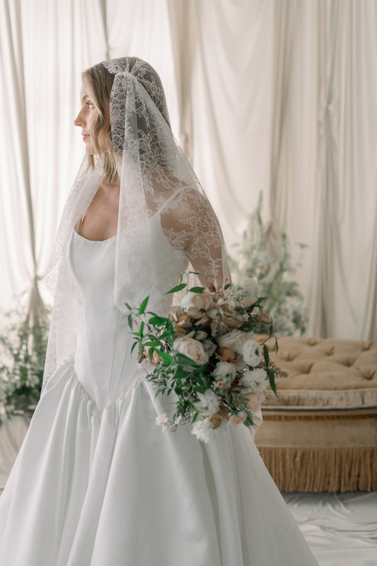 Bride in a white dress holding a bouquet with a lace veil, standing in a softly lit room.