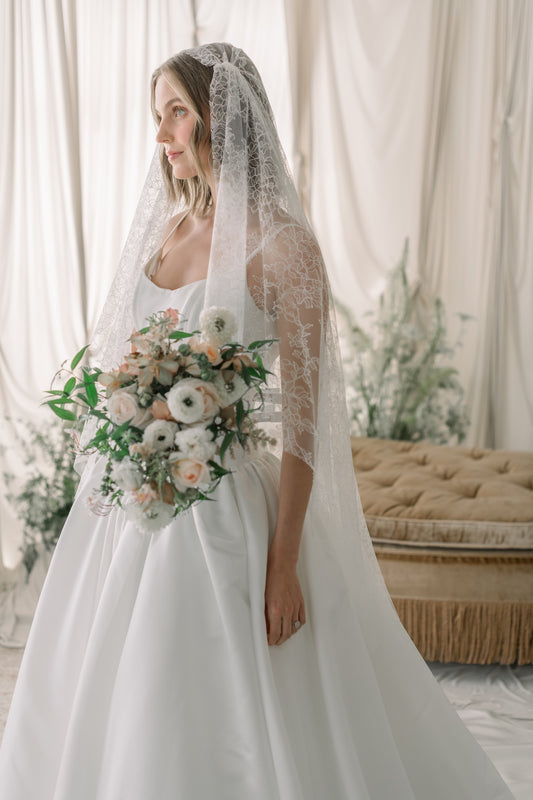 Bride in a white wedding dress with a lace veil holding a bouquet in a decorated room.