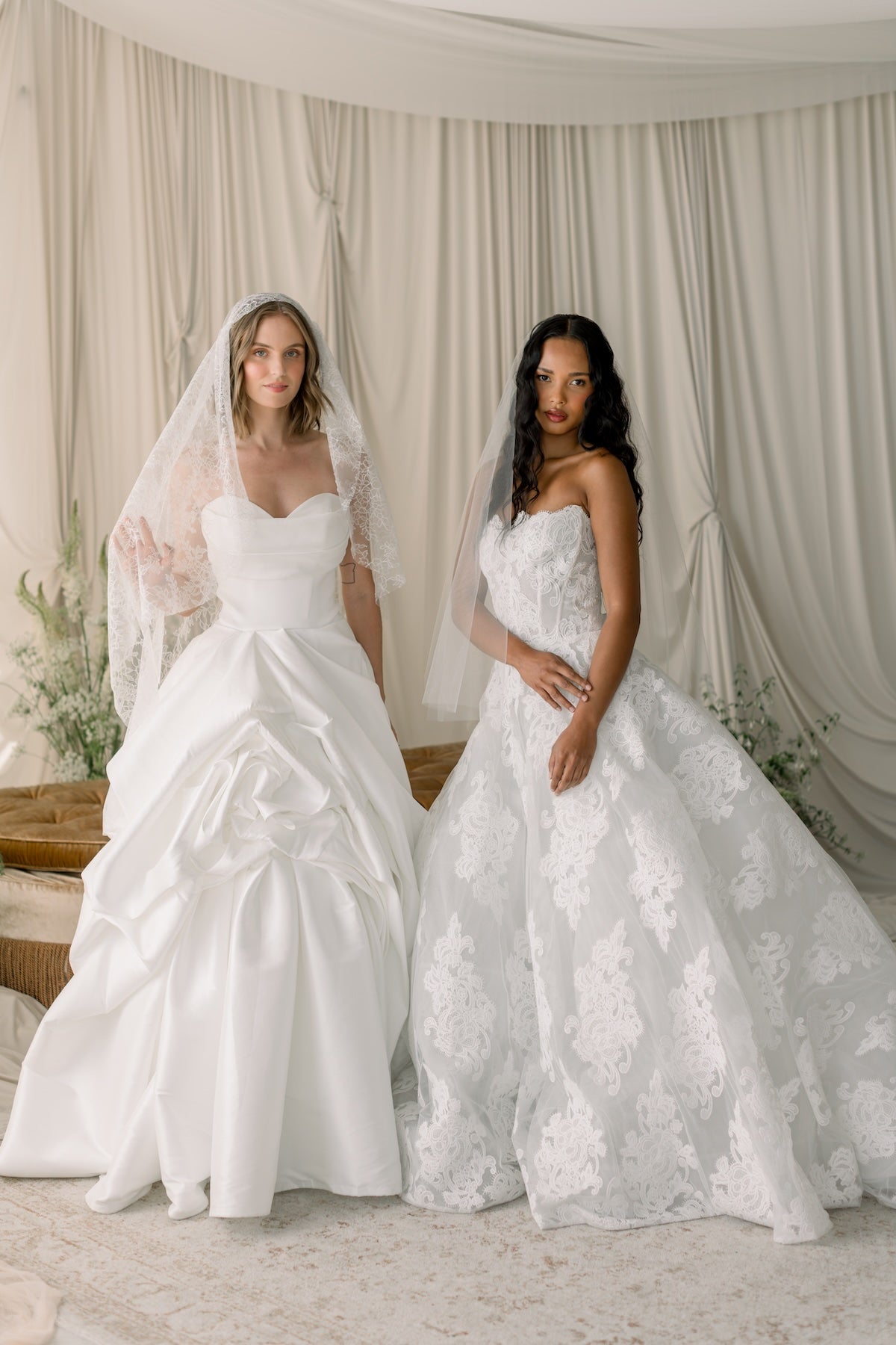Two women in elegant white wedding dresses standing against a neutral background