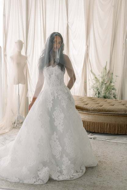 Woman wearing a white lace wedding dress in a softly lit room with curtains and a bench.