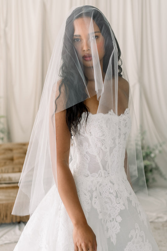 Bride wearing a white lace wedding dress with a sheer veil against a neutral background