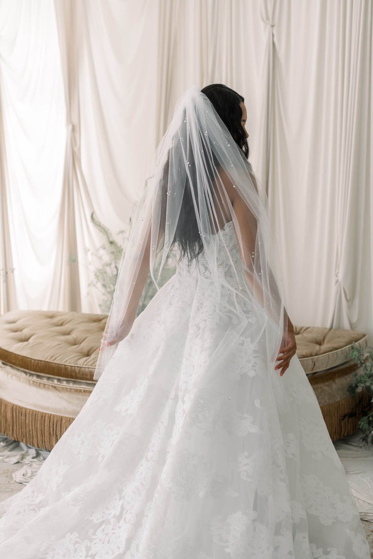 Bride in a white wedding dress with a long veil standing in front of white curtains.