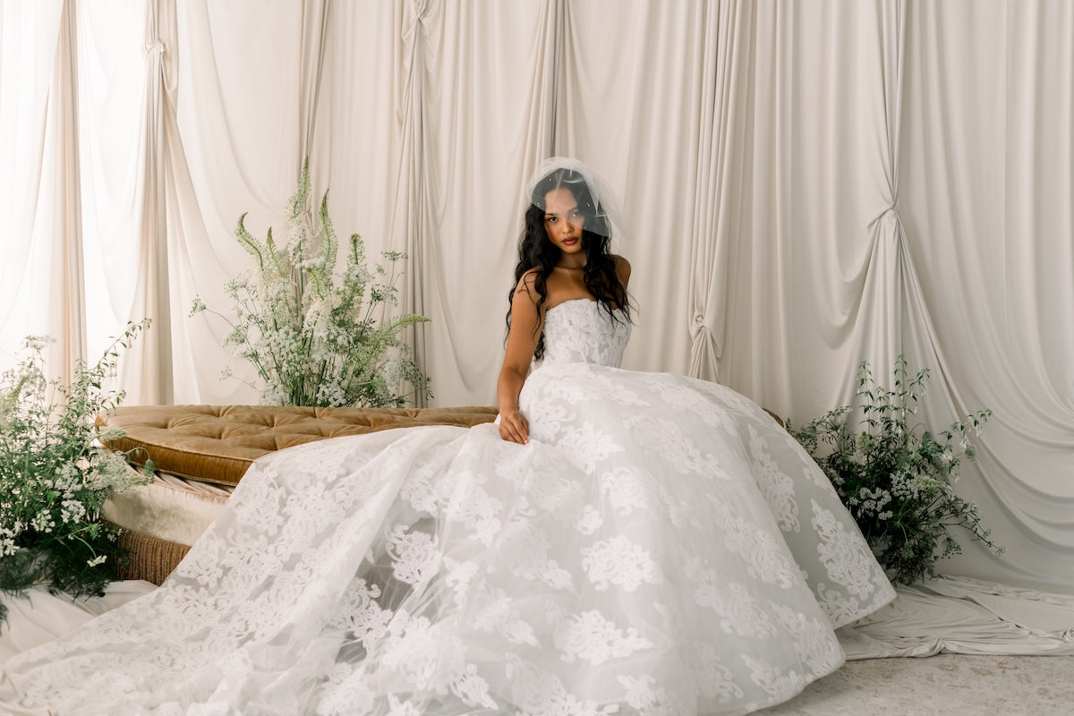Woman in a white wedding dress with floral details sitting on a bed with white curtains and flowers in the background.