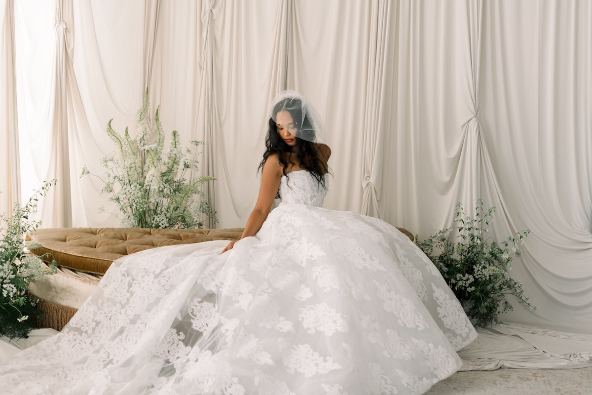 Woman in a white wedding dress sitting on a couch with floral decorations and white curtains in the background.